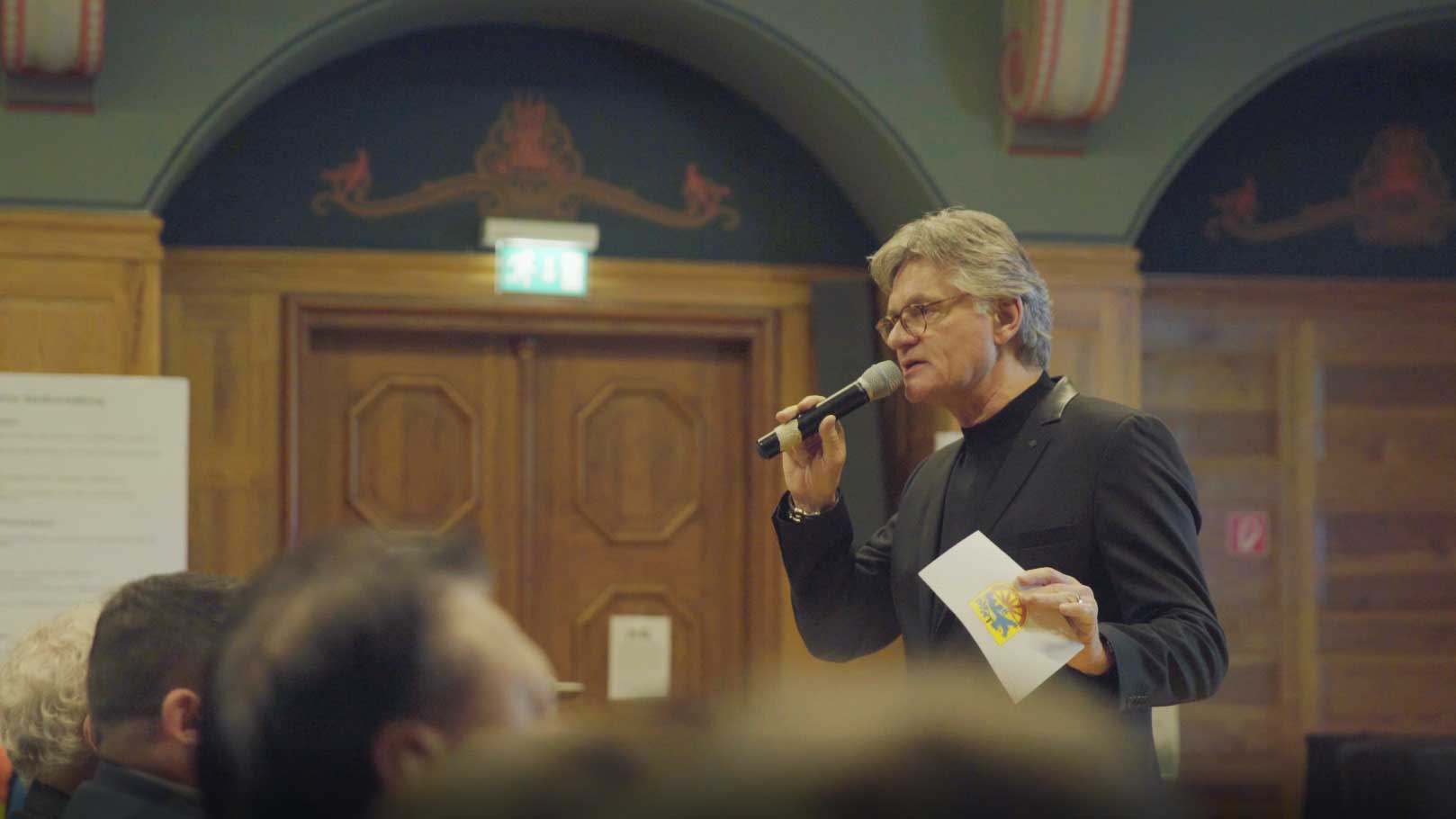 Peter Escher steht mit Mikrofon im Publikum in der Aula